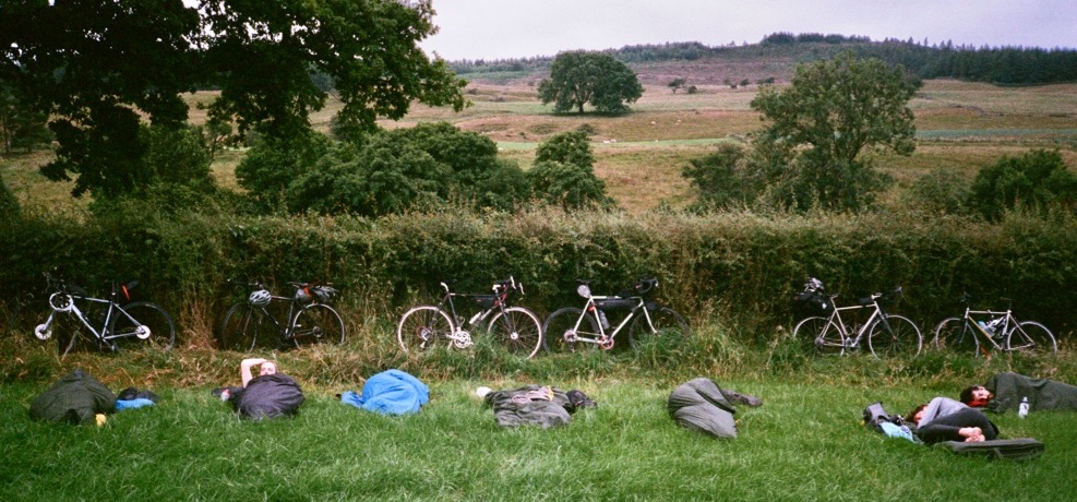 Six people sleeping in bivvy bags in a field. There are bikes propped against a hedge in the backgroud.